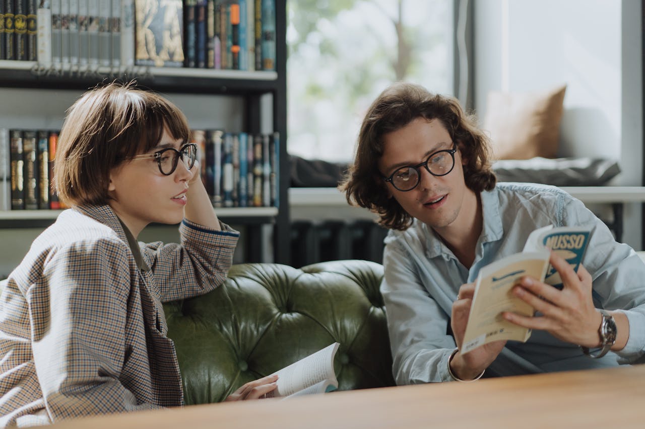 Two young adults in a library discussing books on a comfortable green sofa.